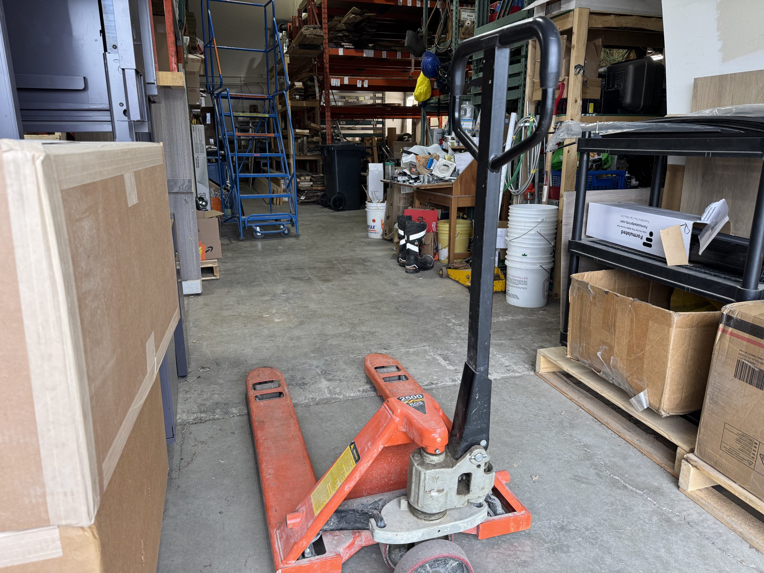 Pallet jack in the foreground of the shared warehouse floor at The Corner Coworking in Cochrane, with pallet racks and member inventory in the background